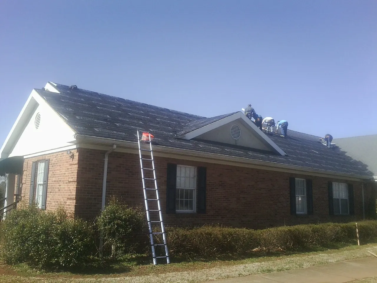 Crew tearing shingles off a brick ranch roof with a ladder set — Metal Roof Repair work by our crew in Swanzey