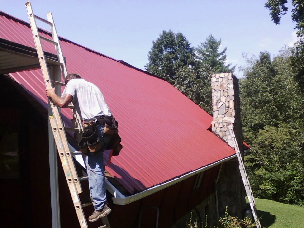 Roofer on a ladder beside a red metal roof and stone chimney — Soffit & Fascia Repair by Roofing Brothers in Swanzey