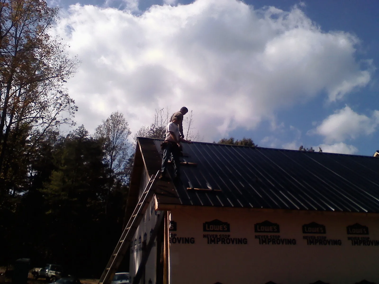 Roofer fastening dark metal panels high against a cloudy sky, from a Roof Inspection job in Swanzey